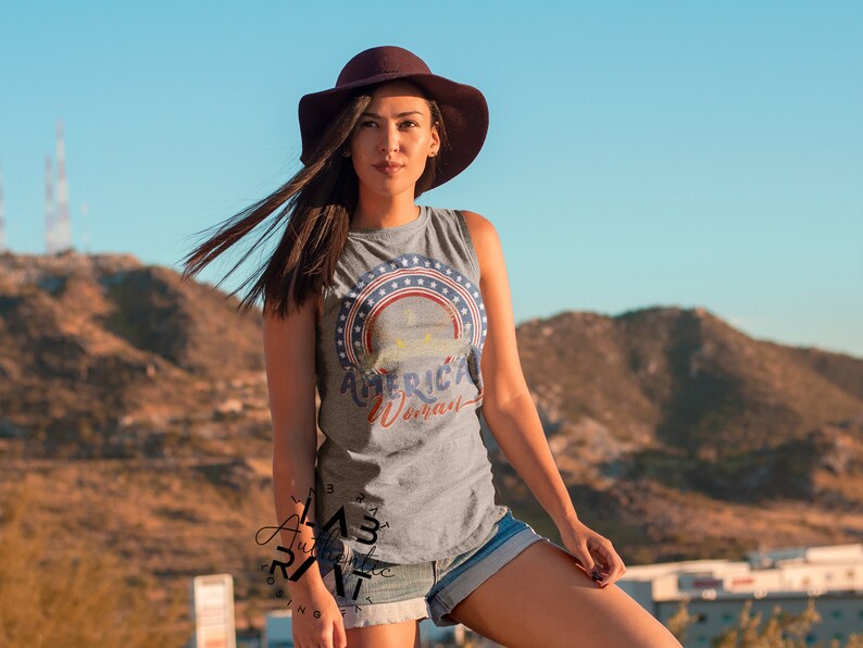 May include: A woman wearing a grey sleeveless shirt with a red, white, and blue graphic that says "American Woman". She is wearing a brown hat and denim shorts. The background is a blurry image of a mountain.