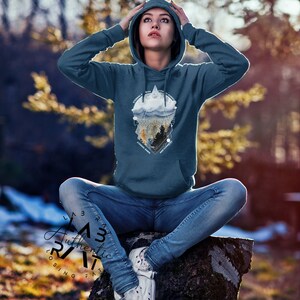 May include: A teal blue hoodie with a graphic print of a mountain range, clouds, and trees. The graphic is in a geometric shape with the text "Explore" in the center. The hoodie is worn by a person sitting on a tree stump.