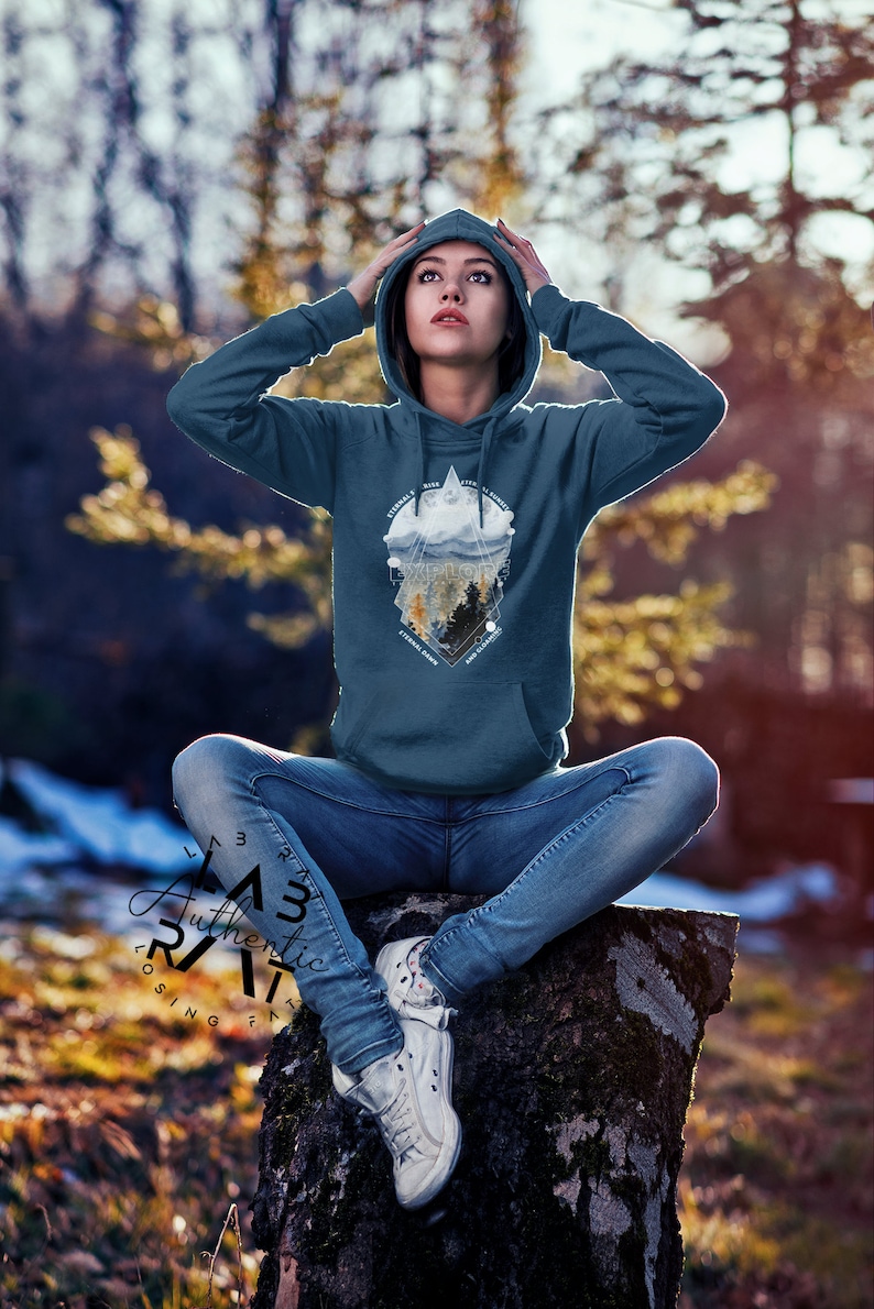 May include: A teal blue hoodie with a graphic print of a mountain range, clouds, and trees. The graphic is in a geometric shape with the text "Explore" in the center. The hoodie is worn by a person sitting on a tree stump.