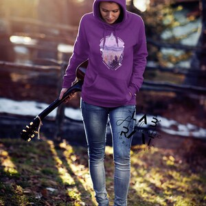 May include: A woman wearing a purple hoodie with a geometric design featuring a mountain landscape and the word "Explore". The hoodie has a drawstring hood and kangaroo pocket. She is holding a guitar and walking on a path in a forest.