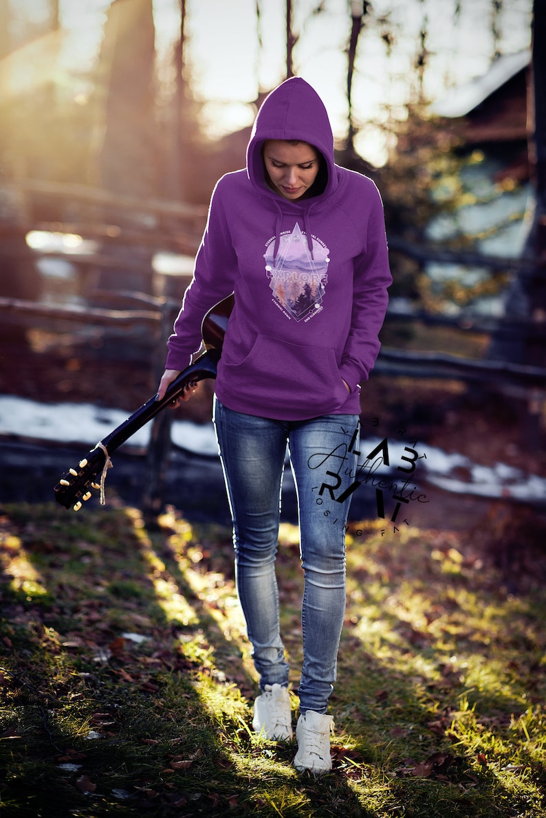 May include: A woman wearing a purple hoodie with a geometric design featuring a mountain landscape and the word "Explore". The hoodie has a drawstring hood and kangaroo pocket. She is holding a guitar and walking on a path in a forest.