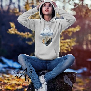 May include: A gray hooded sweatshirt with a graphic print of a mountain range and trees in a geometric shape. The text "Explore" is printed in the center of the graphic. The model is wearing the sweatshirt and sitting on a tree stump.