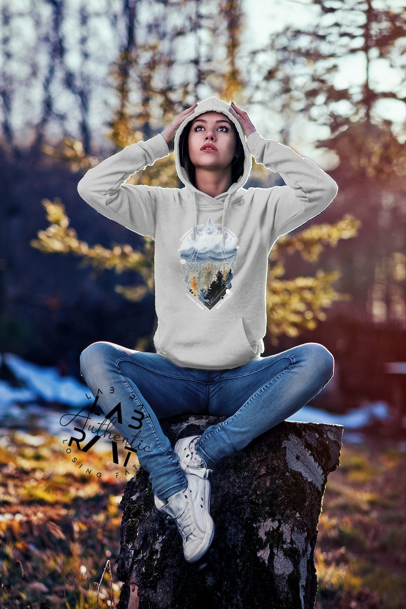 May include: A gray hooded sweatshirt with a graphic print of a mountain range and trees in a geometric shape. The text "Explore" is printed in the center of the graphic. The model is wearing the sweatshirt and sitting on a tree stump.
