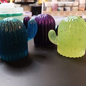 May include: Three resin cactus figurines in shades of blue, purple, and green, each with glitter accents. The cactus shapes have arms and are arranged on a dark surface. The figurines are decorative and whimsical.