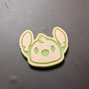 May include: A light green and beige character-shaped craft. The character has large ears, a round face, and three small tufts of hair. The outline and details are in light green, with a beige interior. The craft is likely a decorative item or embellishment.