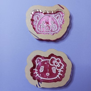 May include: Two craft molds featuring cartoon characters. The top mold has a red panda design with white outlines, set within a light brown, cloud-shaped frame. The bottom mold showcases a Hello Kitty design with white outlines, also in a cloud-shaped frame.