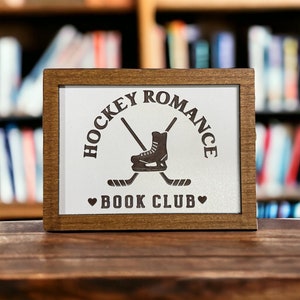 May include: A wooden sign with a white background and brown text. The sign says "Hockey Romance Book Club" with a hockey skate and crossed hockey sticks.
