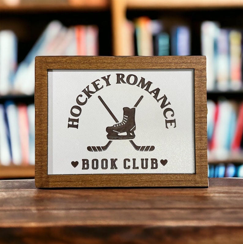 May include: A wooden sign with a white background and brown text. The sign says "Hockey Romance Book Club" with a hockey skate and crossed hockey sticks.