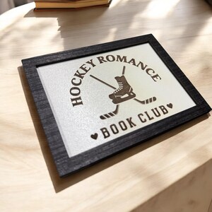 May include: A black framed sign with white background and brown text that reads "Hockey Romance Book Club". A hockey skate with crossed sticks is in the center of the sign.