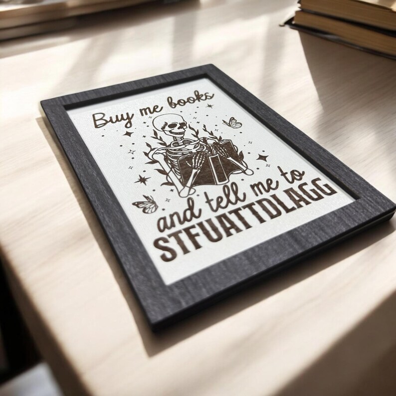 May include: A framed print with a black frame and a white background. The print features a skeleton reading a book, with the text "Buy me books and tell me to STFUATTDLAGG". Decorative butterflies and stars surround the skeleton.