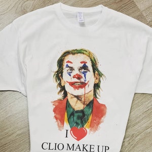 Può includere: Maglietta bianca con un'illustrazione ad acquerello del Joker del film Joker. Il Joker indossa un abito rosso e verde e ha un sorriso rosso dipinto sul viso. Il testo "I ♥ CLIO MAKE UP" è stampato sotto l'immagine.
