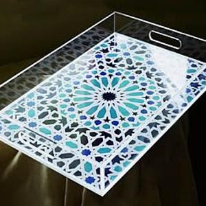 Op de afbeelding: Een transparante acryl serveerschaal met een geometrisch patroon in tinten blauw, groen en wit. De schaal heeft twee handvatten aan de zijkanten.