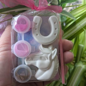 Puede incluir: Un kit de manualidades en una bolsa de pl&aacute;stico transparente con un lazo rosa, que contiene una herradura de yeso blanco, una cabeza de caballo de yeso blanco, tres recipientes circulares de pintura rosa y un pincel fino rosa sobre cart&oacute;n.