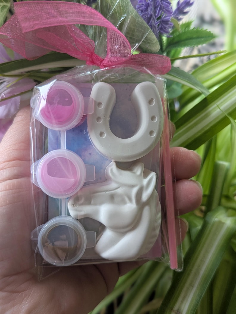 Puede incluir: Un kit de manualidades en una bolsa de pl&aacute;stico transparente con un lazo rosa, que contiene una herradura de yeso blanco, una cabeza de caballo de yeso blanco, tres recipientes circulares de pintura rosa y un pincel fino rosa sobre cart&oacute;n.