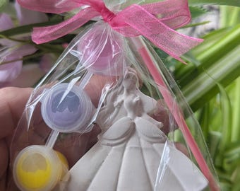 Könnte beinhalten: Ein klarer Cellophanbeutel, gebunden mit einem rosa Band, enthält eine weiße Prinzessinnenfigur, zwei Farbtöpfe mit gelber und violetter Farbe und einen rosa Pinsel. Der Hintergrund umfasst grünes und violettes Laub.