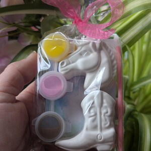 Puede incluir: Una bolsa de pl&aacute;stico transparente atada con una cinta rosa que contiene una figura de cabeza de caballo de cer&aacute;mica blanca, una bota de vaquero de cer&aacute;mica blanca, un pincel peque&ntilde;o y tres botes de pintura amarilla, rosa y marr&oacute;n.