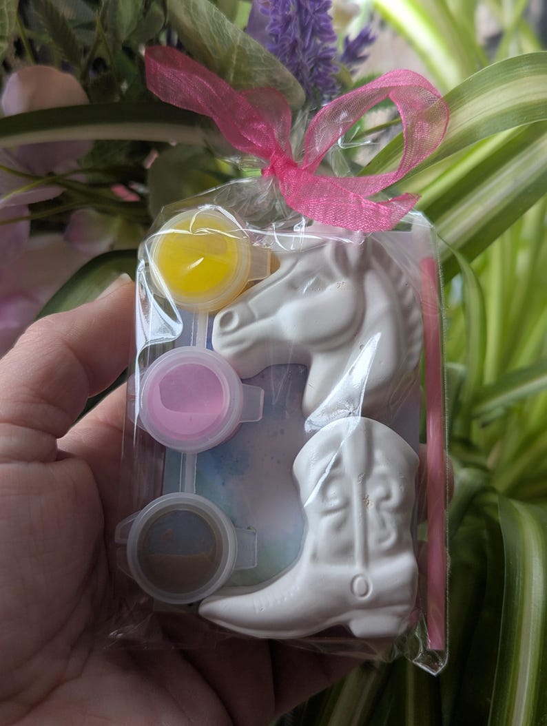 Puede incluir: Una bolsa de pl&aacute;stico transparente atada con una cinta rosa que contiene una figura de cabeza de caballo de cer&aacute;mica blanca, una bota de vaquero de cer&aacute;mica blanca, un pincel peque&ntilde;o y tres botes de pintura amarilla, rosa y marr&oacute;n.