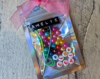 Puede incluir: Una bolsa transparente iridiscente atada con una cinta rosa, que contiene cuentas de colores y cuerdas elásticas. La bolsa tiene una etiqueta que dice "AMELIE" y cuentas que deletrean "ELEA".