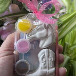 Puede incluir: Una bolsa de pl&aacute;stico transparente atada con una cinta rosa que contiene dos figuras de yeso blanco, una con forma de cabeza de caballo y otra de bota de vaquero, junto con tres peque&ntilde;os botes de pintura redondos en amarillo, rosa y marr&oacute;n.