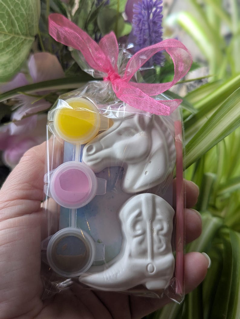 Puede incluir: Una bolsa de pl&aacute;stico transparente atada con una cinta rosa que contiene dos figuras de yeso blanco, una con forma de cabeza de caballo y otra de bota de vaquero, junto con tres peque&ntilde;os botes de pintura redondos en amarillo, rosa y marr&oacute;n.