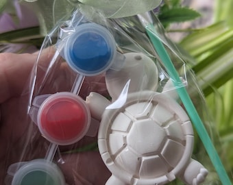 Könnte beinhalten: Ein klarer Zellophanbeutel, gebunden mit einem hellgrünen Band, enthält eine weiße Schildkrötenfigur und drei kleine Farbtöpfe mit blauer, roter und grüner Farbe. Ein grüner Pinsel ist ebenfalls enthalten. Der Hintergrund ist mit Grün verschwommen.