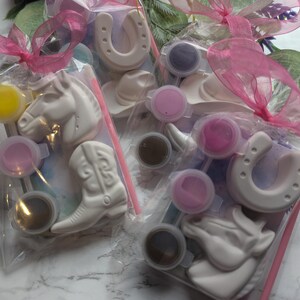 Puede incluir: Tres bolsas de pl&aacute;stico transparente que contienen figuras de yeso blanco con forma de caballos, botas de vaquero y herraduras. Cada bolsa incluye peque&ntilde;os recipientes circulares de pintura de varios colores y un pincel fino, atados con una cinta rosa.
