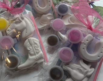 Könnte beinhalten: Drei durchsichtige Plastiktüten mit weißen Gipsfiguren in Form von Pferden, Cowboystiefeln und Hufeisen. Jede Tüte enthält kleine runde Farbbehälter in verschiedenen Farben und einen dünnen Pinsel, zusammengebunden mit einem rosa Schleifenband.