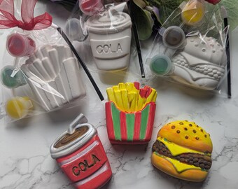 Könnte beinhalten: Eine Sammlung von Keramikfiguren zum Selbstbemalen, darunter ein Burger, Pommes Frites und ein Cola-Becher. Jeder weiße Artikel wird mit kleinen Farbtöpfen geliefert. Der Burger hat gelben Käse und einen braunen Fleischbelag.