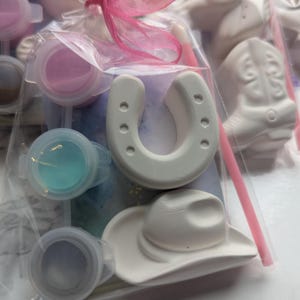Puede incluir: Una bolsa de pl&aacute;stico transparente que contiene figuras de yeso blanco con forma de herradura y sombrero de vaquero, junto con tres peque&ntilde;os botes de pintura redondos en rosa, azul y gris, un pincel rosa fino y un lazo rosa atado arriba.