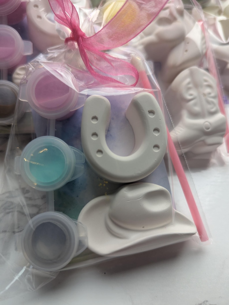 Puede incluir: Una bolsa de pl&aacute;stico transparente que contiene figuras de yeso blanco con forma de herradura y sombrero de vaquero, junto con tres peque&ntilde;os botes de pintura redondos en rosa, azul y gris, un pincel rosa fino y un lazo rosa atado arriba.