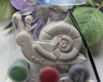 Könnte beinhalten: Ein weißes Keramik-Schneckenfiguren-Malset, verpackt in durchsichtigem Zellophan und mit einem hellblauen Band gebunden. Das Set enthält drei kleine Farbtöpfe in Grün, Rot und Grau sowie einen Pinsel. Die Schnecke hat ein detailliertes Gehäusedesign.
