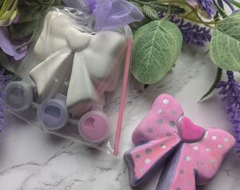 Könnte beinhalten: Ein Mal-dein-eigenes-Schleifen-Set. Das Set enthält eine weiße Keramikschleife, Farbtöpfe in Rosa- und Lilatönen und einen rosa Strohhalm. Eine bemalte rosa Schleife mit silbernen Punkten und einem Herz ist ebenfalls abgebildet.