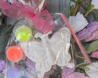 Könnte beinhalten: Ein klarer Zellophanbeutel, der mit einem rosa Band gebunden ist, enthält ein weißes, schmetterlingsförmiges Bastelobjekt, Farbtöpfe in Orange, Lila und Gelbgrün sowie einen rosa Pinsel. Der Hintergrund zeigt rosa und lila Blumen.