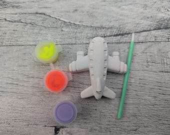 Könnte beinhalten: Ein weißes Flugzeug-Bastelset mit drei kleinen Farbtöpfen in Gelb, Orange und Lila. Ein grüner Pinsel ist ebenfalls enthalten. Die Artikel sind auf einer grauen Holzoberfläche angeordnet.