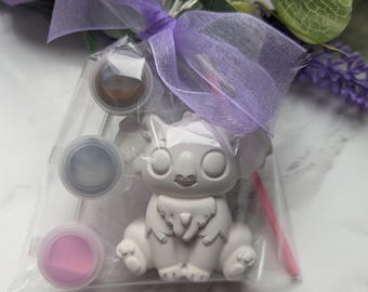 Könnte beinhalten: Ein Mal-dein-eigenes-Koala-Bären-Set in einer transparenten Zellophantüte, gebunden mit einem lavendelfarbenen Band. Das Set enthält eine weiße Koala-Figur, drei Farbtöpfe in Braun, Grau und Rosa sowie einen rosa Pinsel. Dekorative lila Blumen sind im Hintergrund.