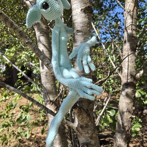 May include: A light blue crocheted elf doll with large black eyes, long arms, and pointed ears, perched on a tree branch. The handmade doll has a slender body and is made of soft yarn.