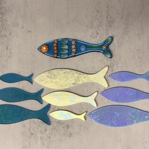 May include: A collection of decorative fish in various sizes and colors. The top fish is blue with orange and green accents. Other fish are shades of blue, yellow, and purple. The fish are arranged on a textured, neutral background.