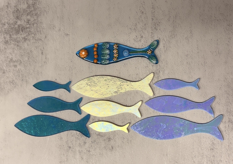 May include: A collection of decorative fish in various sizes and colors. The top fish is blue with orange and green accents. Other fish are shades of blue, yellow, and purple. The fish are arranged on a textured, neutral background.