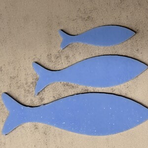 May include: Three blue fish cutouts of varying sizes are arranged on a textured, light brown surface. The fish shapes have a speckled appearance and are outlined in black. The arrangement suggests a decorative or artistic theme.