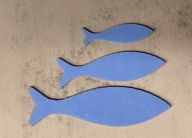May include: Three blue fish cutouts of varying sizes are arranged on a textured, light brown surface. The fish shapes have a speckled appearance and are outlined in black. The arrangement suggests a decorative or artistic theme.