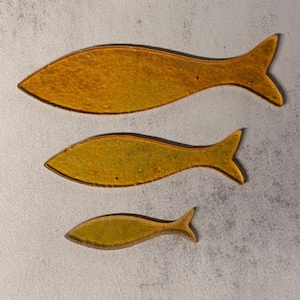 May include: Three decorative fish in varying sizes. The fish are a golden yellow color with a darker brown outline. The fish are arranged vertically, with the largest at the top and the smallest at the bottom.