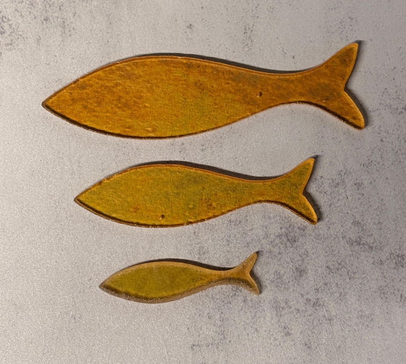 May include: Three decorative fish in varying sizes. The fish are a golden yellow color with a darker brown outline. The fish are arranged vertically, with the largest at the top and the smallest at the bottom.