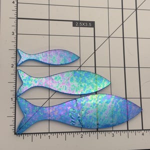 May include: Three iridescent fish-shaped glass pieces in varying sizes. The fish have a blue, purple, and green color scheme. The pieces are on a grid background with measurements in inches.
