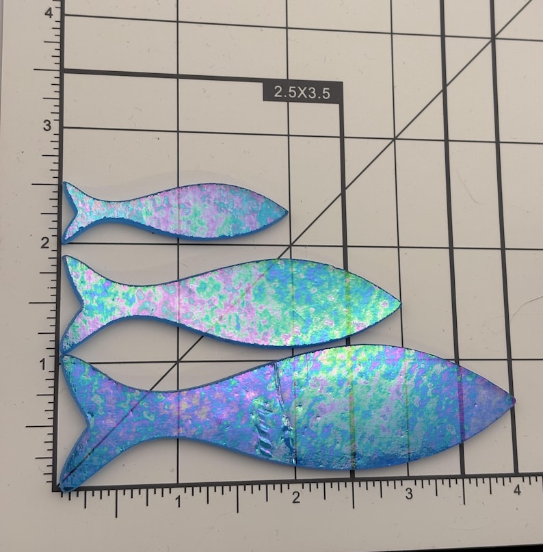 May include: Three iridescent fish-shaped glass pieces in varying sizes. The fish have a blue, purple, and green color scheme. The pieces are on a grid background with measurements in inches.