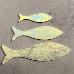 May include: Three fish-shaped decorative objects in varying sizes. The fish are painted in shades of yellow, blue, and gray, with a textured, mottled appearance. The fish are arranged on a gray surface.
