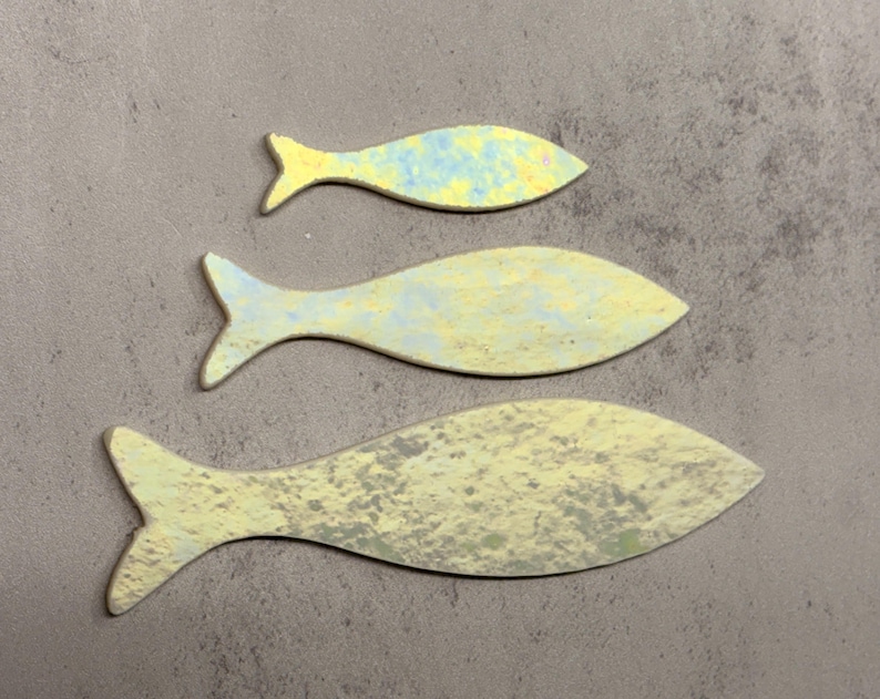 May include: Three fish-shaped decorative objects in varying sizes. The fish are painted in shades of yellow, blue, and gray, with a textured, mottled appearance. The fish are arranged on a gray surface.