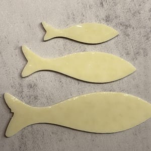 May include: Three light yellow fish-shaped objects of varying sizes are arranged on a gray surface. Each fish has a tail fin and a smooth, glossy appearance. The fish shapes are simple and stylized.