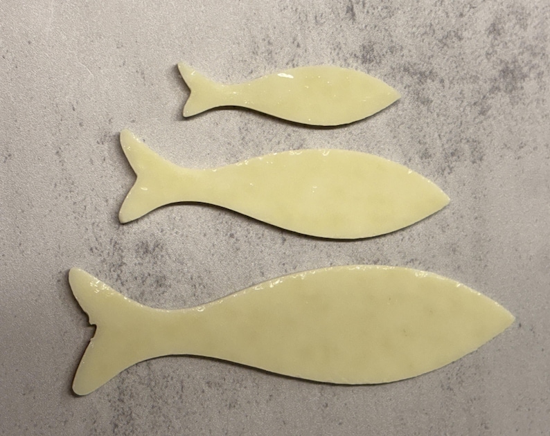 May include: Three light yellow fish-shaped objects of varying sizes are arranged on a gray surface. Each fish has a tail fin and a smooth, glossy appearance. The fish shapes are simple and stylized.