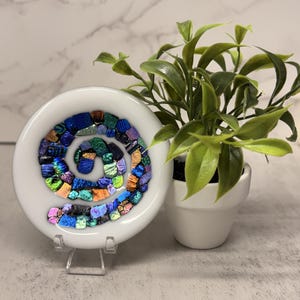 May include: A white, round decorative dish with a spiral design made of colorful, iridescent glass pieces. The dish is displayed on a clear stand, with a small potted plant in the background.