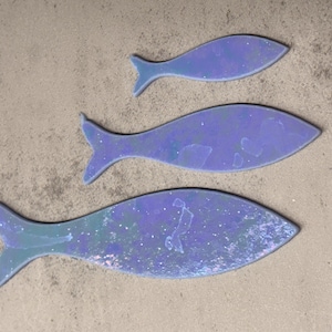 May include: Three iridescent blue fish cutouts of varying sizes are arranged on a textured, light brown surface. The fish have a shimmering, speckled appearance, with a gradient of blue and green hues. The fish tails are clearly defined.